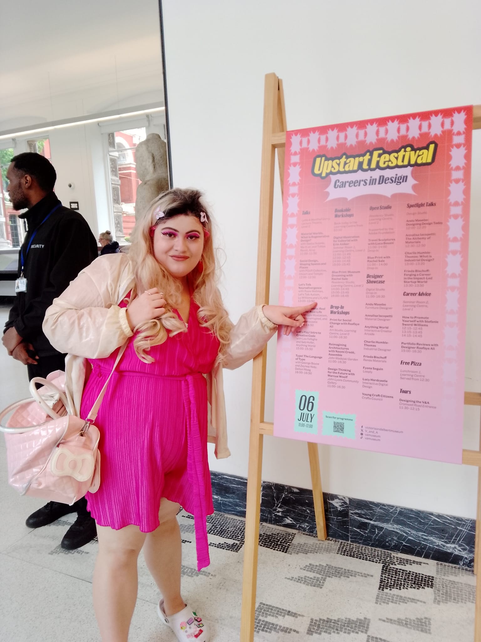 Lylani, a non-binary White femme with a thick build, wearing the biggest blonde wig ever and a pink satin playsuit, points to her name on the poster for the V&A's 2024 Upstart festival