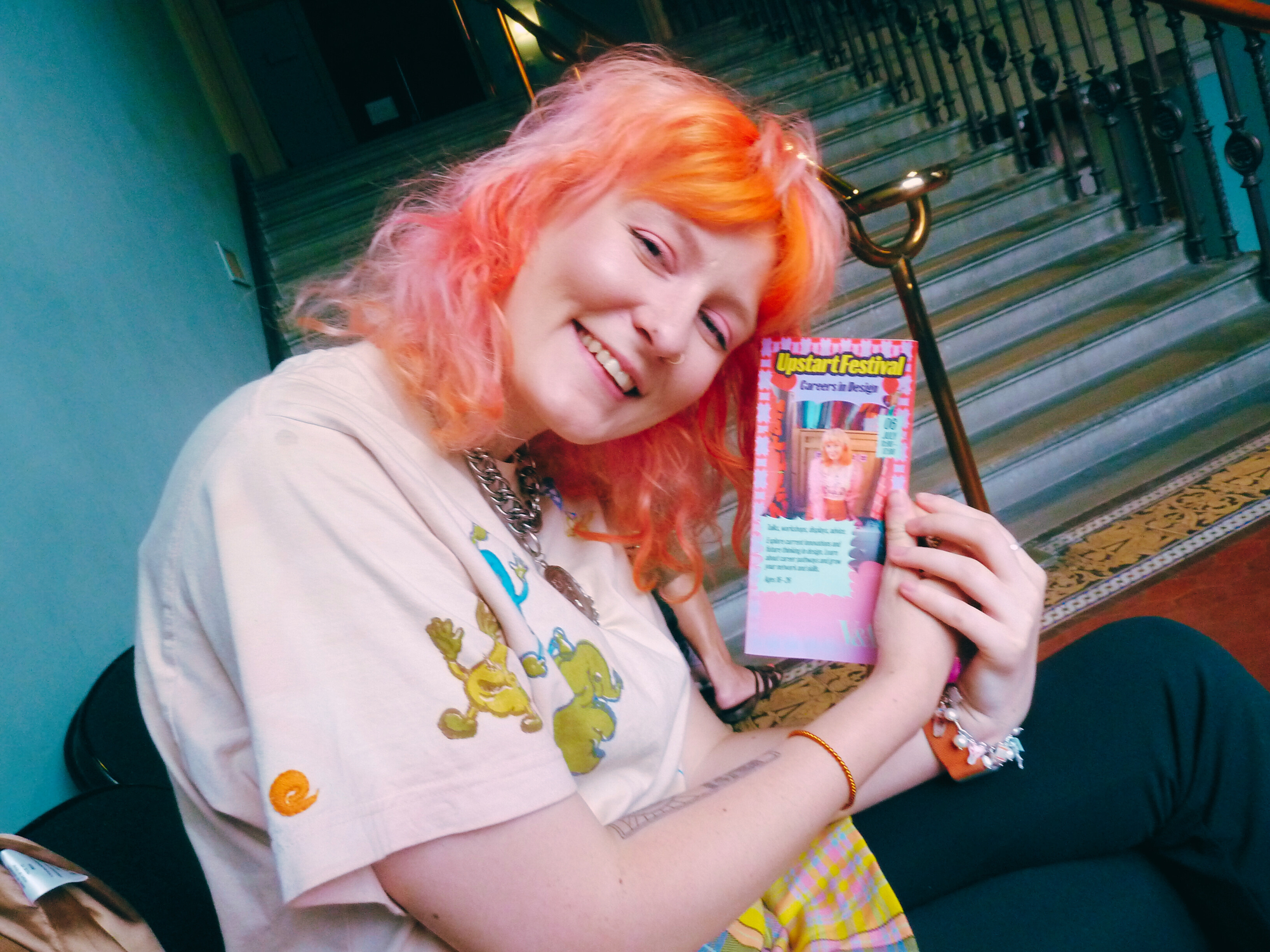 Lu Williams, a White non-binary femme with pink and orange hair, poses with a flyer for the 2024 Upstart festival, of which they are on the cover.